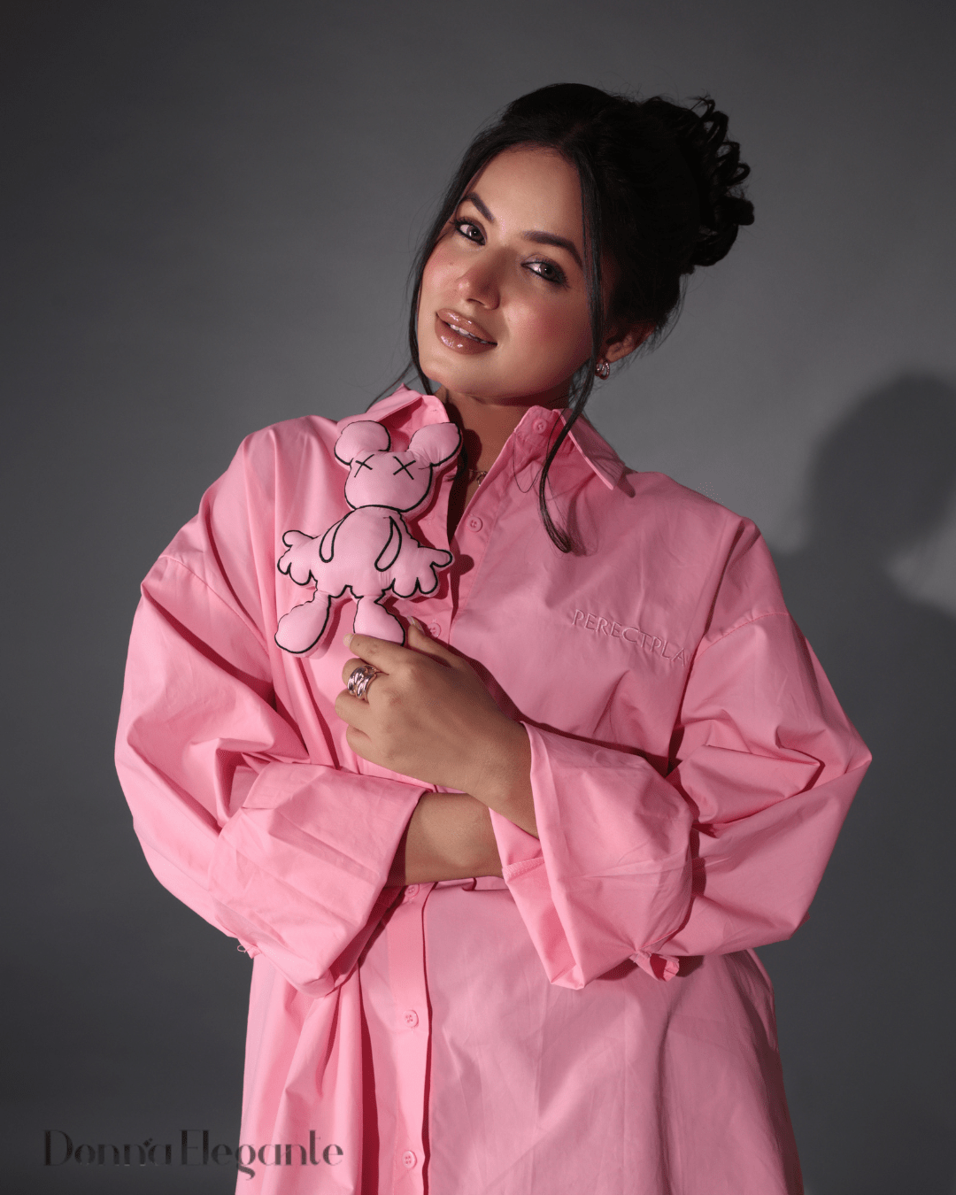 Woman posing in a loose pink shirt dress while holding a matching cartoon plush toy, styled in soft indoor lighting.