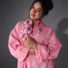 Woman posing in a loose pink shirt dress while holding a matching cartoon plush toy, styled in soft indoor lighting.