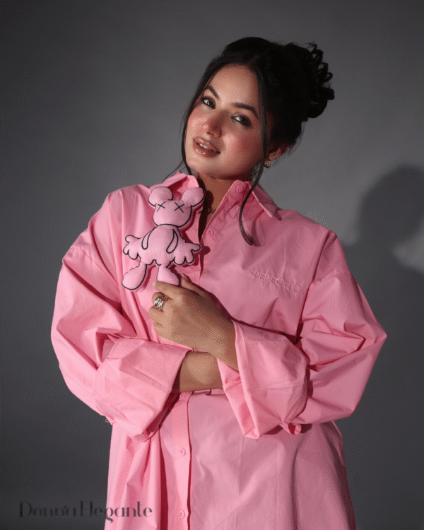 Woman posing in a loose pink shirt dress while holding a matching cartoon plush toy, styled in soft indoor lighting.