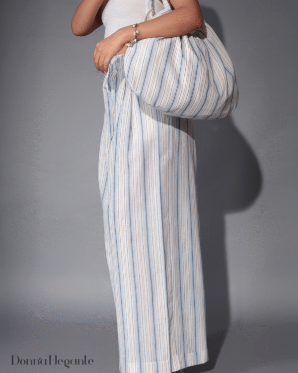 Woman holding a large slouchy striped cotton shoulder bag in neutral and blue tones, styled with matching striped wide-leg pants.